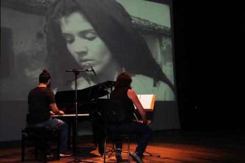 duas pessoas tocando piano e telão com mulher aparecendo ao fundo #paratodosverem