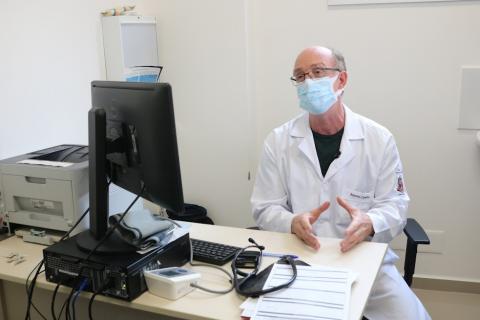 o médico marcos caseiro está sentado em uma cadeira. À mesa à frente dele tem um desktop. #paratodosverem