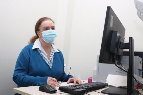 A alergologista e imunologista Luciana Sacconi está sentada à mesa diante de desktop. Ela usa máscara. #paratodosverem