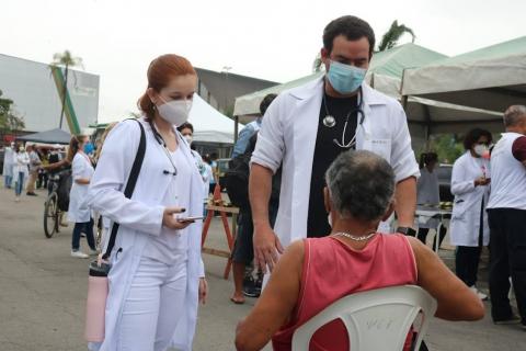 duas pessoas vestidas de branco, com estetoscópio conversam com um homem sentado, de costas para a foto. #paratodosverem