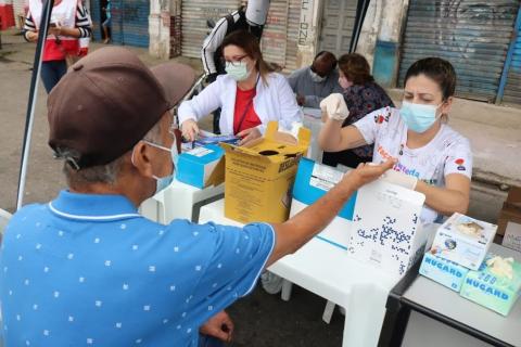 homem está sentado com o braço estendido para uma muher. Ele está fazendo um teste com a retirada de uma gota de sangue do dedo. #paratodosverem