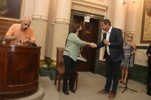O prefeito cumprimenta a homenageada com um aperto de mãos. #paratodosverem