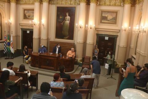 Pessoas sentadas na plateia e na mesa principal. #paratodosverem