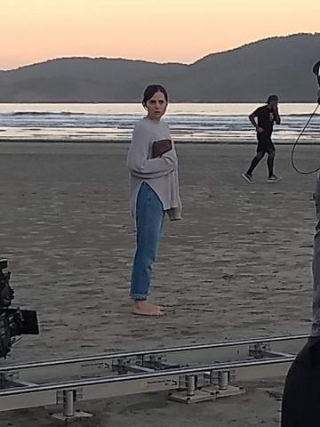Letícia Colin, que faz a personagem Amanda, está na areia da praia, descalça, usando calça e blusa. Ela segura um livro no braço e mão direita. #paratodosverem