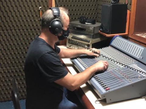Estúdio de gravação digital. Homem manipula ilha de edição. Ele usa fones no ouvido. #paratodosverem