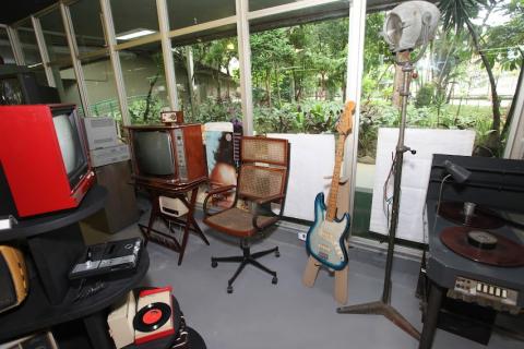 guitarra elétrica, cadeira de balanço, aparelhos de som, TV e outros objetos antigos expostos. #paratodosverem