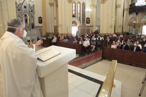 Padre celebra missa. Ele está atrás de púlpito falando ao público que está sentado no templo. #paratodosverem