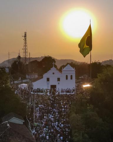 Imagem aérea de drone mostra Santuário de Monte Serrat com fiéis subindo escadaria e por do sol ao fundo. #paratodosverem