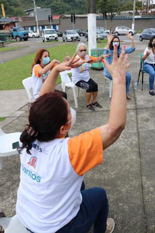 Em primeiro plano, mulher está sentada com os dois braços erguidos. Ao fundo, outras fazem o mesmo movimento. #paratodosverem