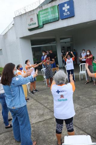 Pessoas em pé fazem movimentos com os braços. Ao fundo, o prédio da policlínica com placas de identificação da unidade, da prefeitura e do SUS. #paratodosverem