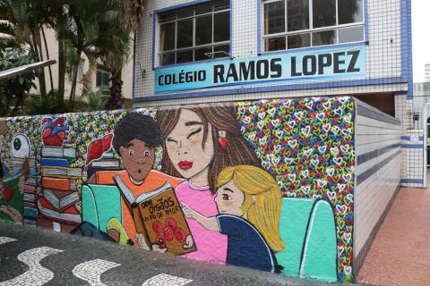 Fachada da escola com placa onde se lê Colégio Ramos Lopez. Abaixo, o mural grafitado com crianças lendo livros. #paratodosverem