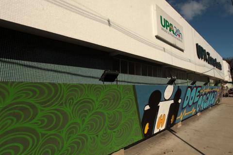 grafite em parede com desenho e texto: doe órgãos. No topo da fachada estão placas da UPA. #paratodosverem