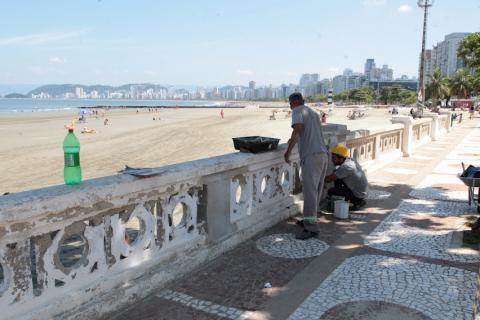 dois homens estão junto a mureta com instrumentos nas mãos. A praia aparece do lado esquerdo. #paratodosverem