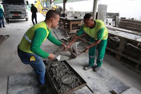 massa é despejada em forma por dois homens . Eles seguram armação de ferro para garantir mais rigidez à peça. #paratodosverem