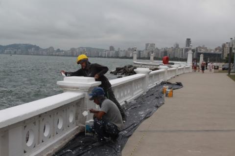 homens pintam mureta diante do mar. O calçadão está à direita. #paratodosverem