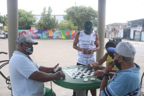 em primeiro plano, homens estão jogando dominó sentados à mesa. Ao fundo está o mural pintado. #paratodosverem