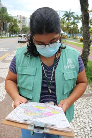 Mulher usando colete de identificação segura e olha para prancheta com mapa de identificação do bairro. #paratodosverem