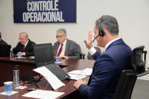 o prefeito rogério santos está de lado para a foto. Ele usa fone e microfone. Ao fundo, outras duas pessoas sentadas. Todos estão com laptops abertos sobre a mesa. #paratodosverem