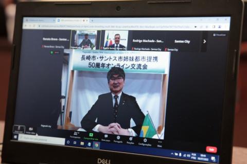 tela de computador com o prefeito de nagasaki ao centro. Ele está com as mãos cruzadas sobre uma mesa. Ao lado direito da imagem há uma pequena bandeira do brasil. #paratodosverem