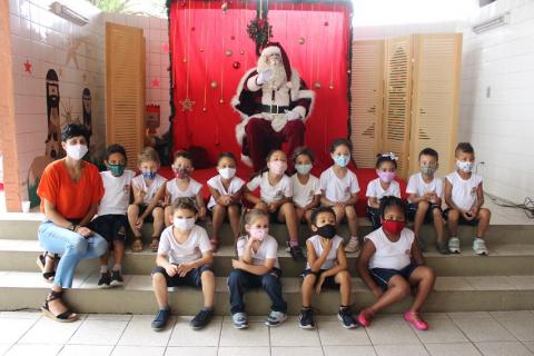 Papai Noel sentado ao fundo com crianças na frente. #paratodosverem