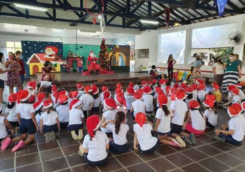 crianças estão sentadas, todas usando gorro de papai noel, assistindo ao papai noel que está no palco. #paratodosverem