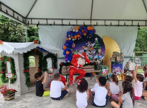 Papai Noel está sentado em banco, ao lado de casinha e conversando com crianças. #paratodosverem