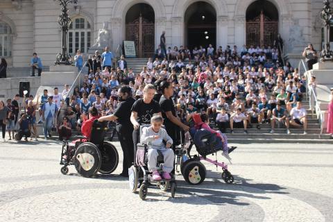 pessoas movimentam cadeirantes em dança e crianças assistem na escaria #paratodosverem