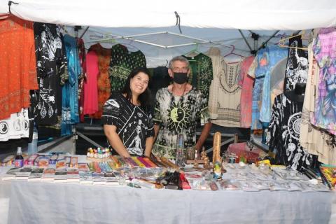 Casal de expositores atrás de mesa com produtos e diante de peças de roupas. #paratodosverem