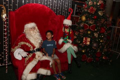 papai noel está sentado com criança ao lado. Mamãe noel acompanha. Há uma árvore de natal à direita. #paratodosverem