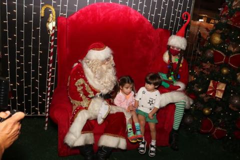 Duas crianças estão sentadas ao lado de papai e mamãe noel. #paratodosverem