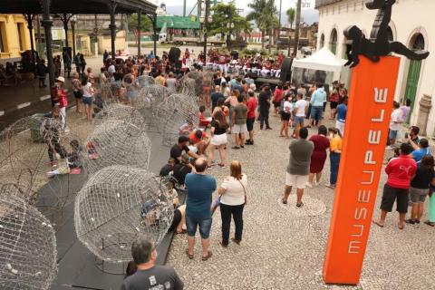 Bolas aramadas de natal enfeitam rua à esquerda. Ao fundo, público assiste apresentação de banda. Em primeiro plano, à direita, o totem do museu pelé. #paratodosverem