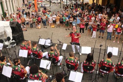 Músico estão em palco diante de suas estantes com partituras. O regente está à frente e o público atrás. #paratodosverem