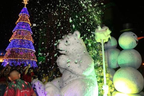 carro alegórico com grande urso polar e bonecos de neve na parte de trás. Do lado esquerdo, uma grande árvore de natal iluminada. O carro lança um efeito de neve no ar. #paratodosverem