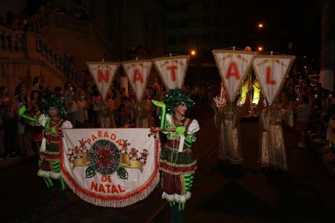 abertura do desfile com personagem segurando uma espécie de faixa onde se lê parada de natal. Atrás deles, cada integrante segura um estandarte com as respectivas letras da palavra natal. #paratodosverem