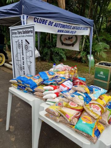 mesa com pacotes de alimentos não perecíveis. Uma tenda ao fundo. #paratodosverem