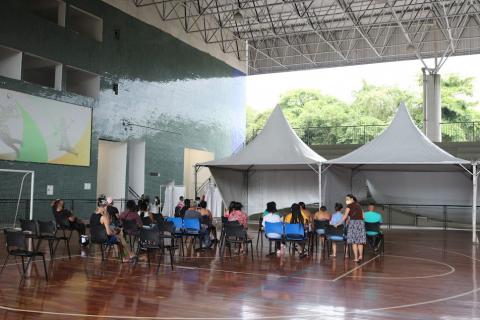 pessoas sentadas em cadeiras diante de duas tendas ao fundo. Ginásio é coberto. #paratodosverem