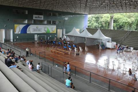 ginásio com arquibancada onde se vê pessoas sentadas. Na quadra estão as cadeiras de uma estrutura de tendas para atendimento. #paratodosverem