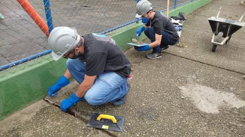 homens usando capacete e luvas fazem reparos no solo e mureta que cerca play ground. Ao lado deles há um carrinho de mão. #paratodosverem