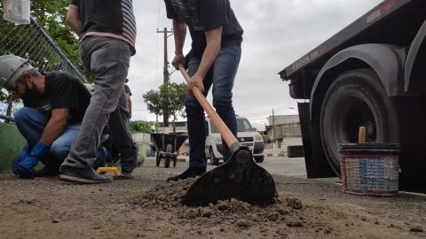 close de pá misturando cimento no chão. Há homens trabalhando por perto, e a roda de uma caminhão estacionado junto à guia, à direita. #paratodosverem 