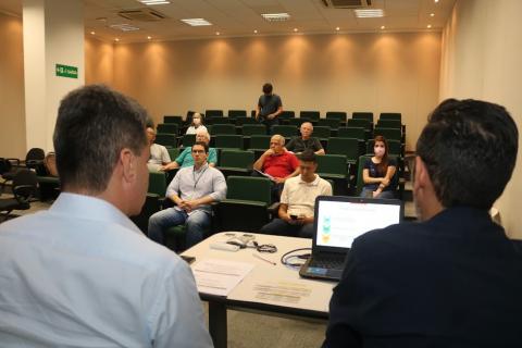 audiência em auditório, com duas pessoas sentadas à frente de um computados em primeiro plano e algumas sentadas ao fundo. #paratodosverem