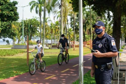 Guarda Municipal observa ciclovia. Ele tem um rádio na mão esquerda. Dois ciclistas passam na ciclovia. #Paratodosverem