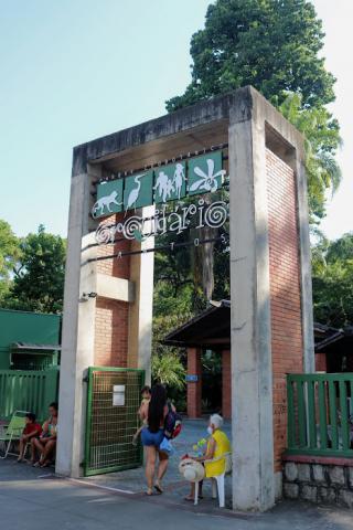 Fachada do Orquidário com um homem sentado à frente e, em pé, mulher entra carregando criança de colo. #pratodosverem  