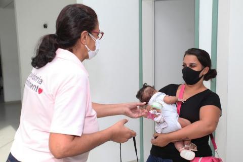 Voluntária usando camiseta cor-de-rosa se aproxima de mãe que segura bebê. #paratodosverem
