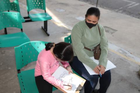 menina faz desenho, sentada, ao lado de uma mulher. #paratodosverem