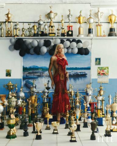 a cantora para foto, usando um vestido longo vermelho. Ela está em uma sala onde estão dispostos vários troféus no chão e prateleiras. Há também um arranjo de balões de gás atrás da artista. #paratodosverem