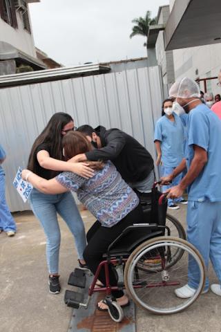 Paciente em cadeira de rodas, na área externa do hospital, abraça familiares. #paratodsoverem