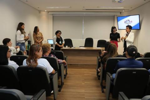 pessoas sentadas em auditório assistem palestra. Há três mulheres em pé ao fundo. #paratodosverem