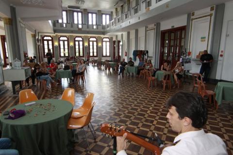 violonista em primeiro plano com salão ao fundo onde estão sentadas mulheres. #paratodosverem