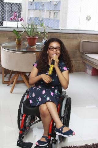 menina na cadeira de rodas segura microfone próximo à boca. #paratodosverem