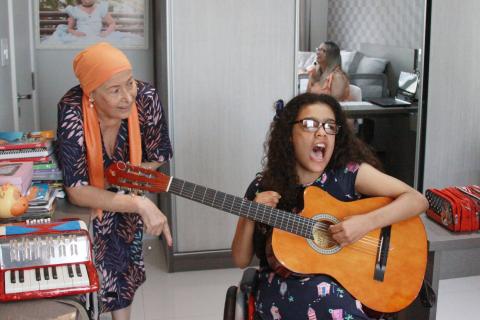 menina toca violão e canta enquanto a mãe a observa. #paratodosverem